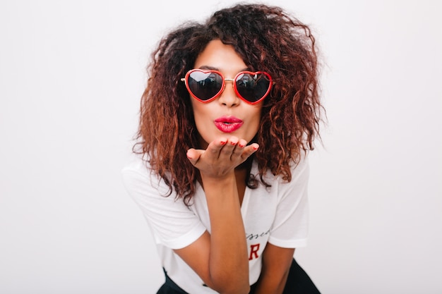 Lovely lady with light-brown skin sending air kiss isolated. Beautiful african female model with curly hairstyle posing in trendy red sunglasses.