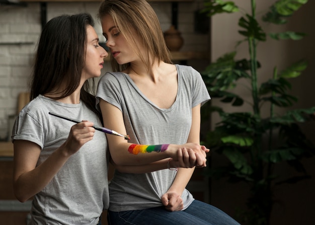 Lovely lesbian woman painting the rainbow flag on her girlfriend's hand with paintbrush