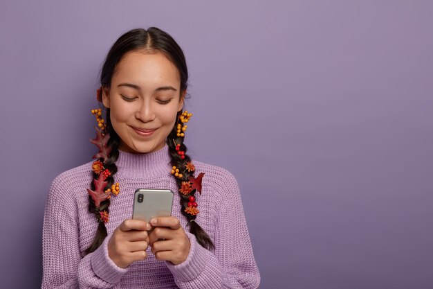 lovely millennial brunette woman has two plaits with stuck autumn leaves as natural cosmetics, focused in smartphone