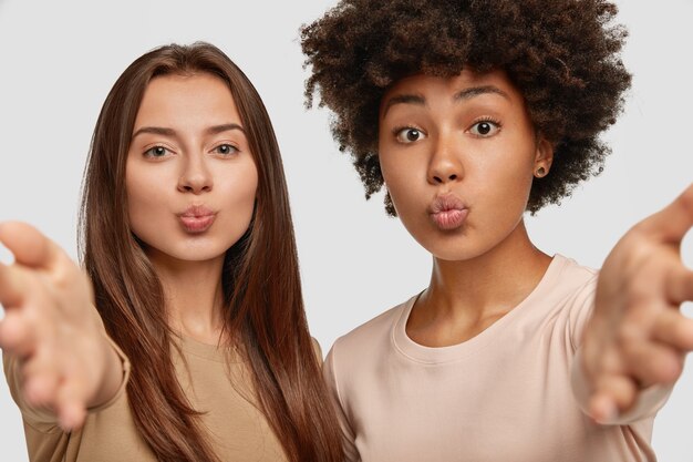 Lovely mixed race women pout lips, stretch hands as going to embrace someone, express their good feeling, pose together against white wall. Interracial female friends indoor.