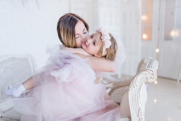 Lovely mom and daughter hug each other tender sitting on white chair