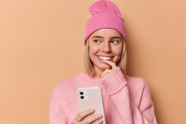 Прекрасная задумчивая европейская женщина использует мобильный телефон, думает о содержимом сообщений, прокручивает социальные сети, радостно смотрит в сторону, носит мягкий свободный розовый джемпер и шляпу, изолированную на коричневом фоне.