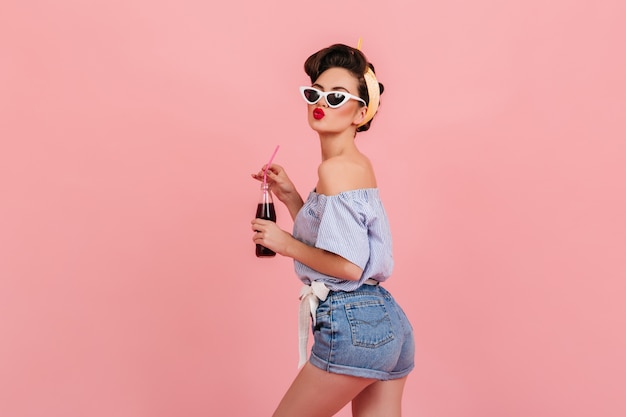 ポジティブな感情を表現するデニムショートパンツの素敵なピンナップガール。ピンクの背景に飲み物とボトルを保持しているサングラスの嬉しいブルネットの女性のスタジオショット。