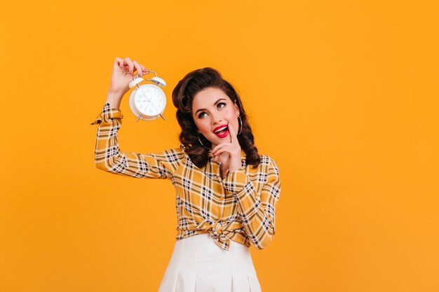 Lovely pinup girl holding big clock. Studio shot of well-dressed european woman isolated on yellow background.