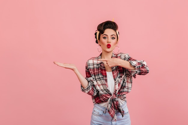 Lovely pinup girl with bright makeup standing on pink background. Studio shot of blissful stylish woman pointing with finger.