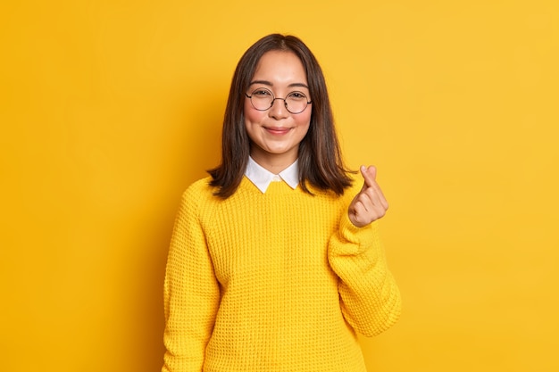 Lovely pleased Asian woman makes korean like gesture smiles gently expresses love to someone