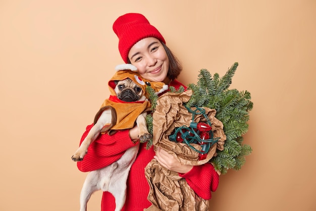 La bella donna bruna contenta posa con il cane carlino e gli attributi delle vacanze si prepara per le celebrazioni di natale o capodanno indossa un maglione rosso e il cappello si gode il tempo festivo con le pose preferite dell'animale domestico al coperto
