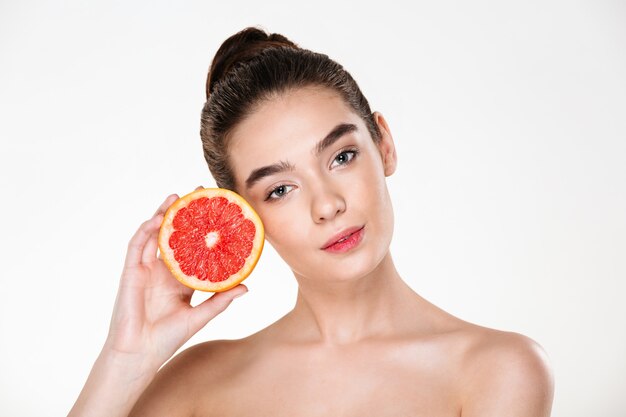 Lovely portrait of pretty half-naked woman with natural makeup holding juicy grapefruit near her face and looking