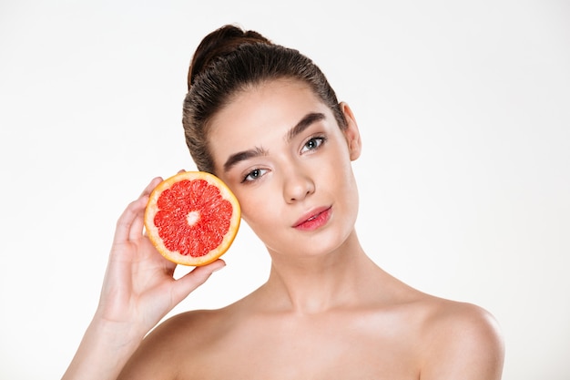 Lovely portrait of pretty half-naked woman with natural makeup holding juicy grapefruit near her face and looking