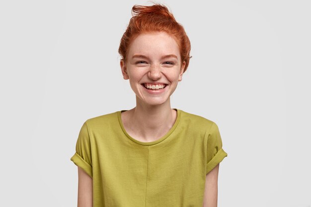 Lovely red haired girl with positive expression, laughs as watches funny TV show, enjoys weekend, dressed in green t shirt, has freckled skin, isolated over white wall, amused by comic idea