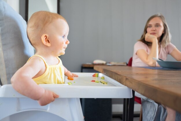 사랑스러운 red-haired 신생아 먹고 흰색 플라스틱 아기 부엌 좌석에 앉아. 나무 테이블에 앉아서 그녀의 아이보고 아름 다운 젊은 어머니. 가족 및 어린 시절 개념