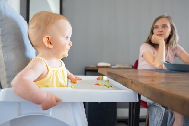 Lovely red-haired newborn eating and sitting in white plastic baby kitchen seat. Beautiful young mother sitting at wooden table and looking at her child. Family and childhood concept