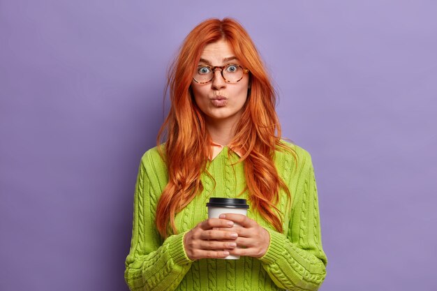 lovely redhaired young woman keeps lips rounded and looks surprisingly holds disposable cup of coffee enjoys spare time wears green jumper.