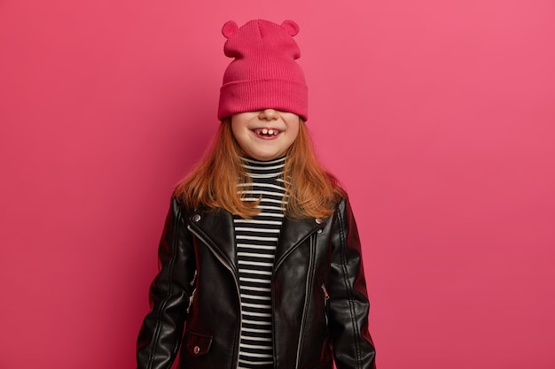 lovely redhead girl plays hide and seek game, waits for surprise with positive emotions, covers eyes with pink hat, wears striped jumper and leather jacket, has fun, poses indoor