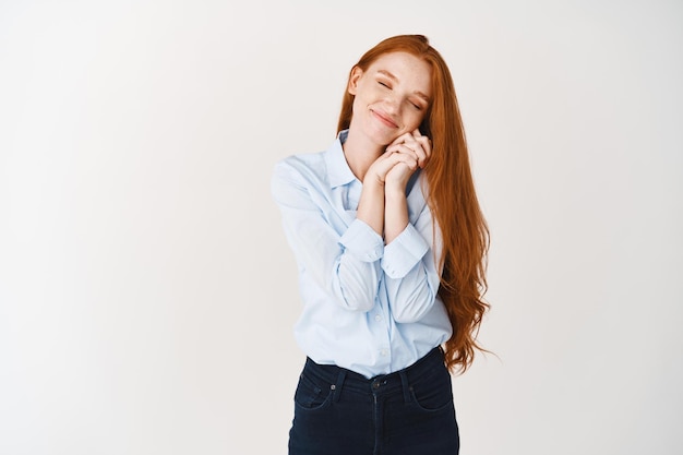 Lovely redhead girl with long ginger hair dreaming, close eyes and leaning on hands, imaging something romantic with heartfelt smile, white wall