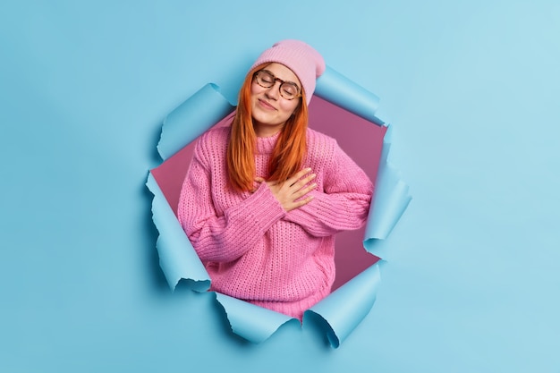 Lovely redhead woman appreciates something makes gratitude gesture closes eyes and stands touched tilts head dressed in pink clothing.