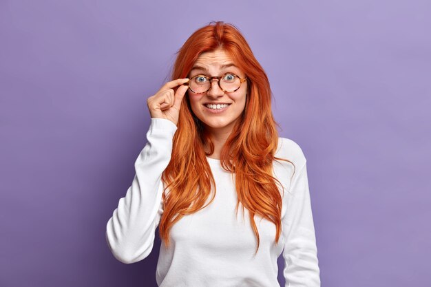 Free photo lovely redhead woman keeps hand on rim of spectacles looks with interest hears something unbelievable wears white casual jumper.