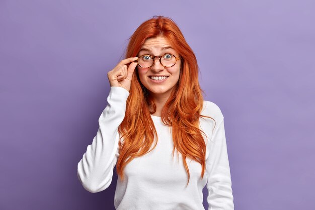 Foto gratuita bella donna rossa tiene la mano sul bordo degli occhiali guarda con interesse sente qualcosa di incredibile indossa un maglione casual bianco.