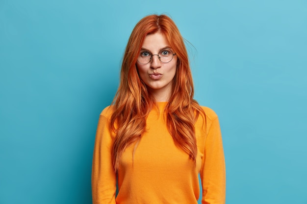 Прекрасная рыжая молодая женщина в круглых очках держит губы сложенными, хочет поцеловать кого-то, одетого в оранжевый джемпер, смотрит прямо.