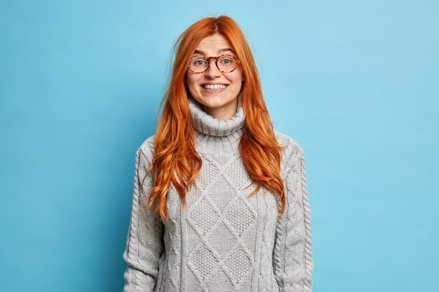 Lovely redhead young woman with european appearance smiles pleasantly dressed in warm sweater expresses positive emotions hears good news.