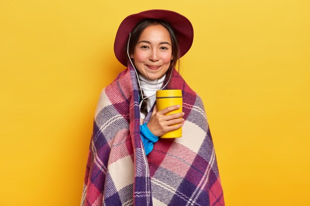 Lovely relaxed female traveler drinks hot beverage from thermos, stands wrapped in plaid, enjoys hiking trip, wears hat, poses over yellow background.