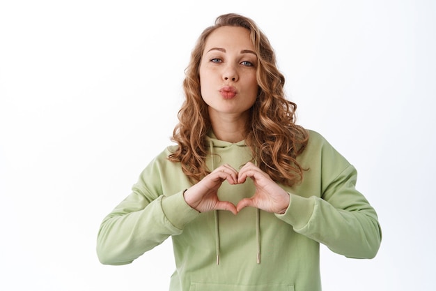 Foto gratuita adorabile ragazza romantica dice ti amo, mostra il segno del cuore e bacia la faccia, in piedi sul muro bianco