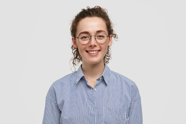 Lovely satisfied freckled female with crisp hair, has gentle smile, dressed in striped black and white shirt, rejoices positive moments in life, isolated over wall. People and emotions