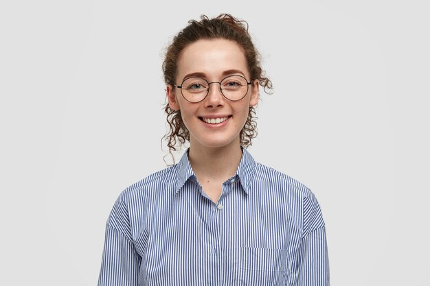 Lovely satisfied freckled female with crisp hair, has gentle smile, dressed in striped black and white shirt, rejoices positive moments in life, isolated over wall. People and emotions