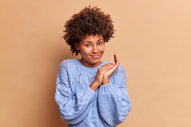 Lovely satisfied woman with Afro hair has satisfied expression rubs palms and smiles gently joyfully wears blue casual jumper sees something very good made up nice plan isolated over brown wall