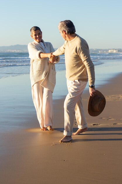 Lovely senior couple having fun at seashore on warm autumn evening. Grey-haired man with hat in hand holding womans hand pulling her forward. Support, relationship, togetherness concept