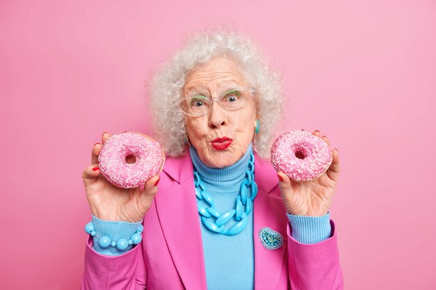 Lovely senior woman holds two glazed donuts keeps lips folded looks pleasantly, dressed in fashionable outfit with necklace and bracelet