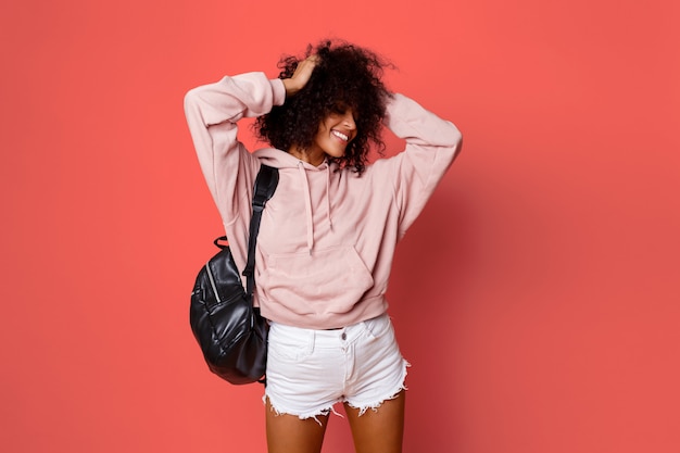 Lovely sexy black woman in stylish hoodie with back pack posing on pink background and playing with curly hairs