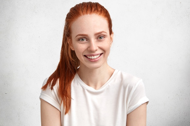 Милая улыбающаяся довольная молодая модель с рыжими волосами и веснушками на лице, одетая в повседневную белую футболку, выражает положительные эмоции
