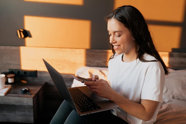 Free photo lovely smiling girl in morning sunlight is working on laptop in the bed happy smiling girl is working at home in bedroom with smartphone and laptop