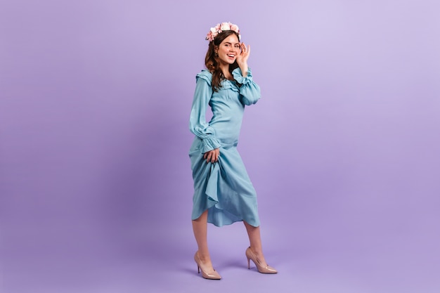 Lovely smiling lady in satin blue outfit. Woman touches her face on lilac wall.