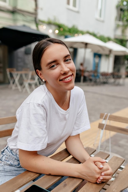 白いTシャツを着て暗い収集された髪の素敵な笑顔の女性は、カフェテリアで屋外で休んでいる間、素晴らしい笑顔でカメラにポーズをとっています