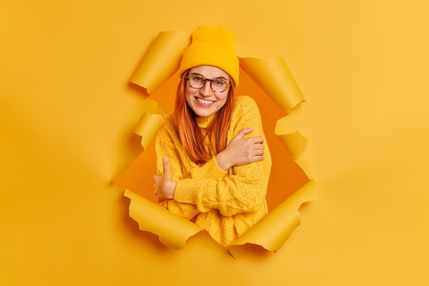 Lovely smiling redhead woman embraces herself feels comfort wears warm knitted sweater looks gladfully.