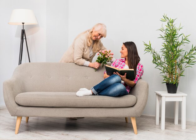 Lovely smiling senior mother giving flower bouquet to her daughter sitting on sofa holding book in hand