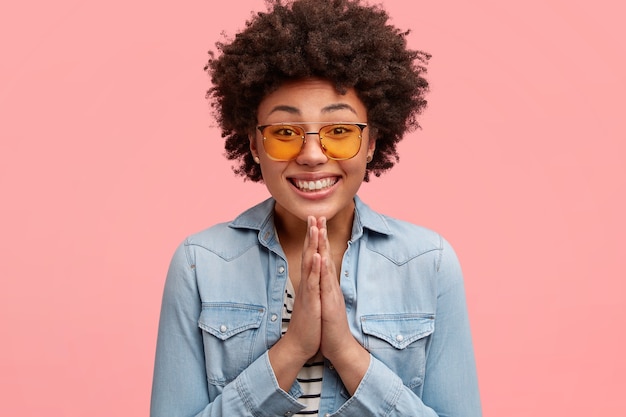 Bella ed elegante giovane donna afroamericana con un ampio sorriso e un'espressione supplichevole, tiene le mani nel gesto di preghiera, chiede qualcosa, indossa una giacca di jeans e occhiali da sole gialli alla moda