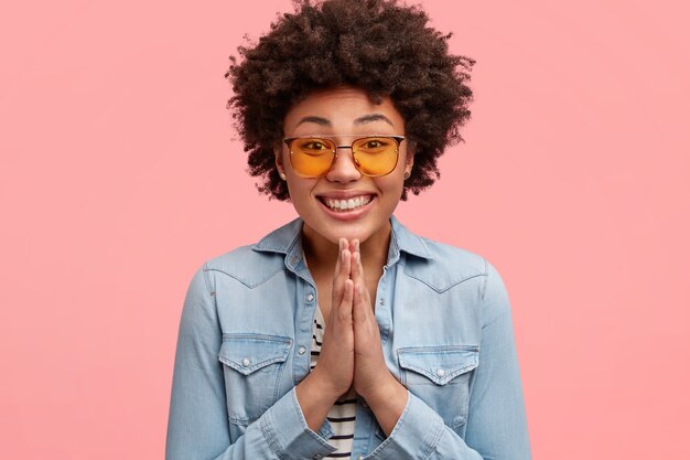 Lovely stylish young African American female with broad smile and pleading expression, keeps hands in praying gesture, asks for something, wears denim jacket and fashionable yellow sunglasses