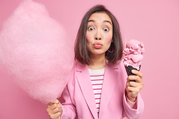 Lovely surprised Asian woman stares  doesnt keep to diet or count calories holds candy floss and big cone ice cream in waffle affordes herself eating sugary treatment during day off