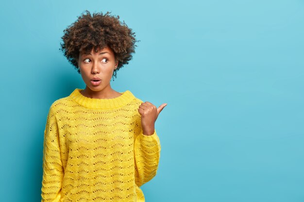 Lovely surprised woman shocked to hear unexpected relevation points thumb away and wears casual jumper impressed by incredible thing poses against blue wall