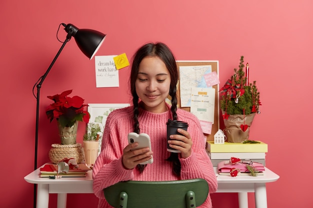 Free photo lovely teenage girl with two plaits holds mobile phone, holds disposable cup of drink