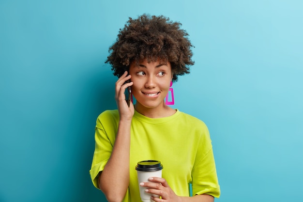 Lovely thoughtful dark skinned woman has telephone conversation concentrated aside pensively bites lips drinks takeaway coffee