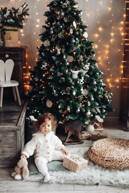 Lovely toddler with a plush pet and a gift sitting on the carpet near Christmas tree