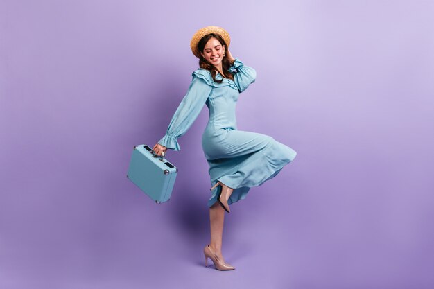 シルクで作られたミディドレスを着た素敵な旅行者は、紫色の壁に楽しくポーズをとっています。スーツケースと麦わら帽子の女の子のフルレングスのショット。