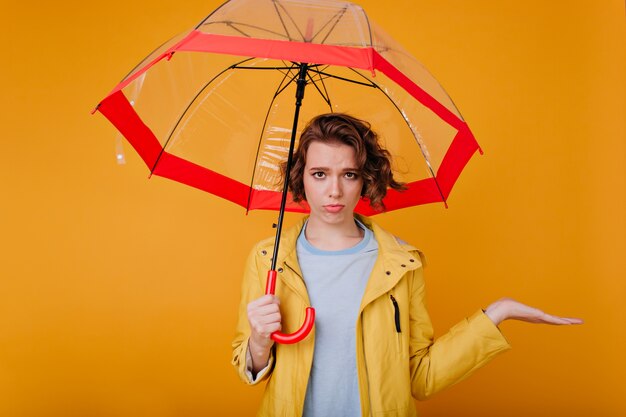 Lovely white girl expressing sad emotions while standing under umbrella. Indoor photo of shy upset lady wears autumn attire holding parasol.