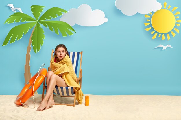 Lovely woman feels cold after swimming in sea, sits at deck chair near palm tree