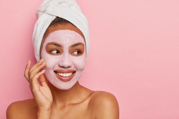 Lovely woman has delicate sooth skin, wears cream mask on face to reduce acnes, has healthy complexion, hygienic treatments wears white wrapped towel on head