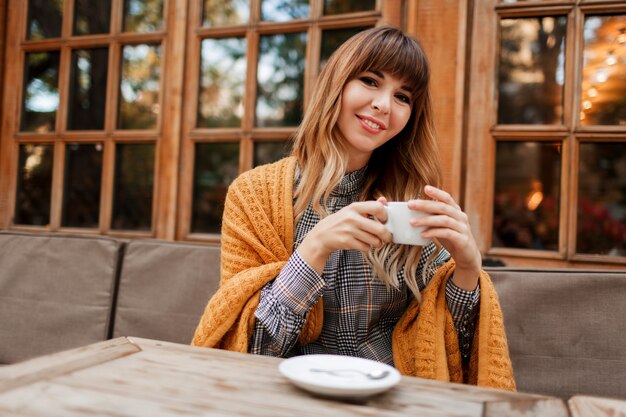 사랑스러운 여자는 나무 인테리어와 아늑한 카페에서 커피 브레이크를 가지고 휴대 전화로 이야기합니다. 뜨거운 카푸치노 한잔 들고. 겨울 시즌. 우아한 드레스와 노란색 체크 무늬를 입고.